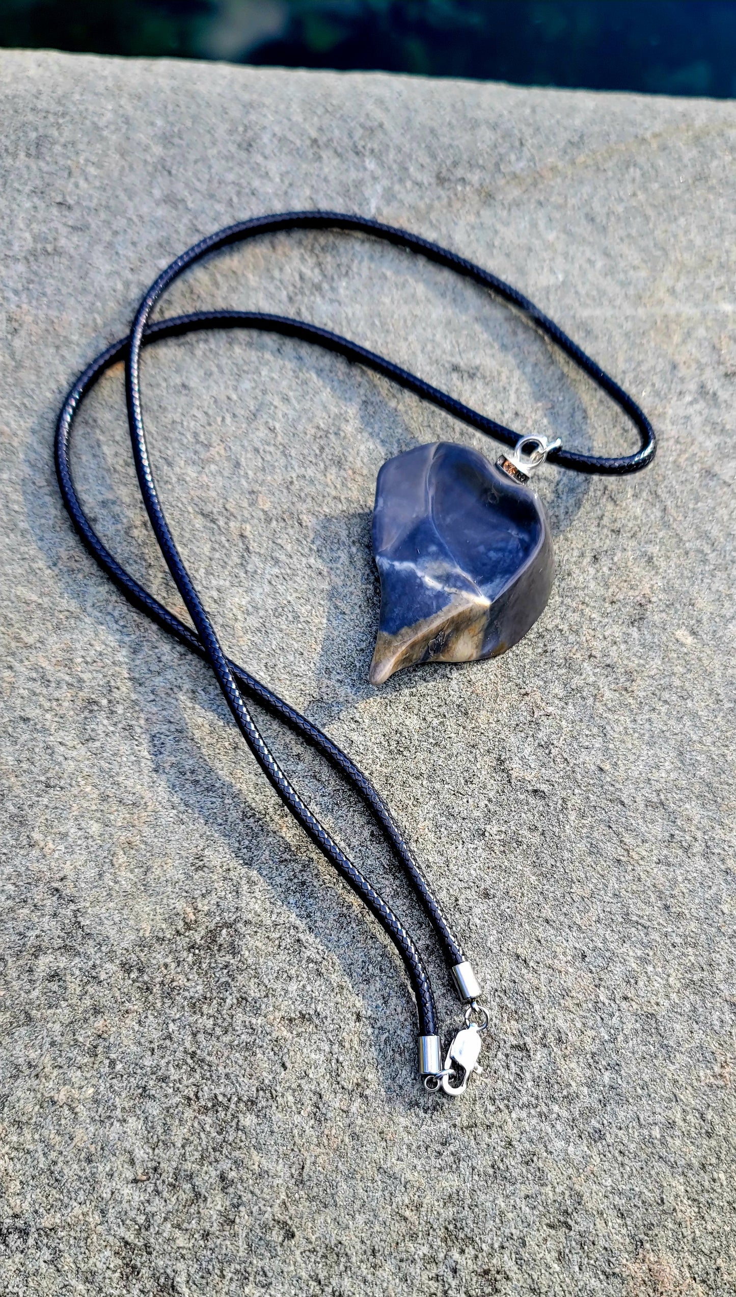 This Polychrome Jasper pendant is sure to be one of the most unique pieces you'll ever come across. Grey and White in color, it has the shape of the continent, Africa. It has stainless steel trim and black string. Pictured hanging over water.