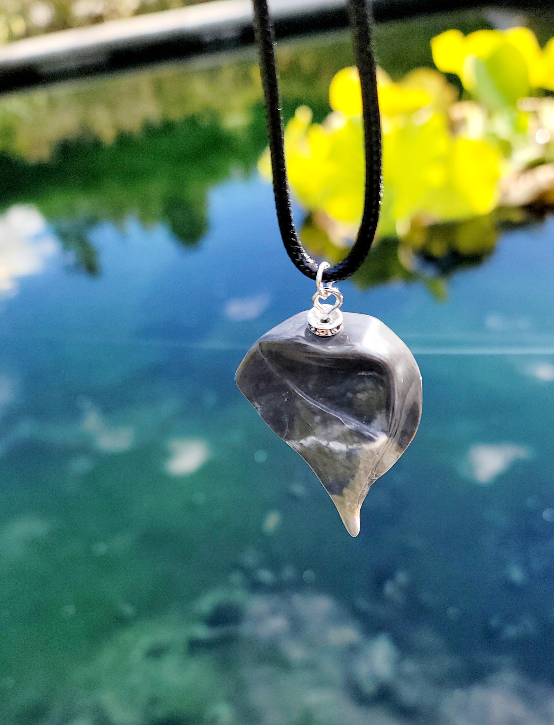 This Polychrome Jasper pendant is sure to be one of the most unique pieces you'll ever come across. Grey and White in color, it has the shape of the continent, Africa. It has stainless steel trim and black string. Pictured hanging over water.