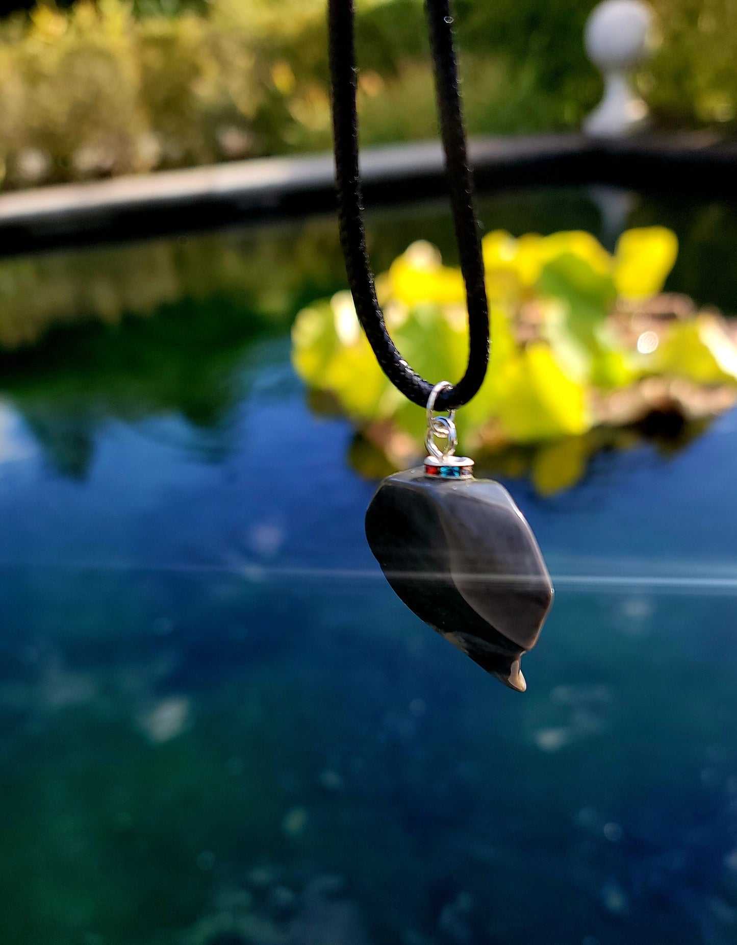 This Polychrome Jasper pendant is sure to be one of the most unique pieces you'll ever come across. Grey and White in color, it has the shape of the continent, Africa. It has stainless steel trim and black string. Pictured hanging over water.