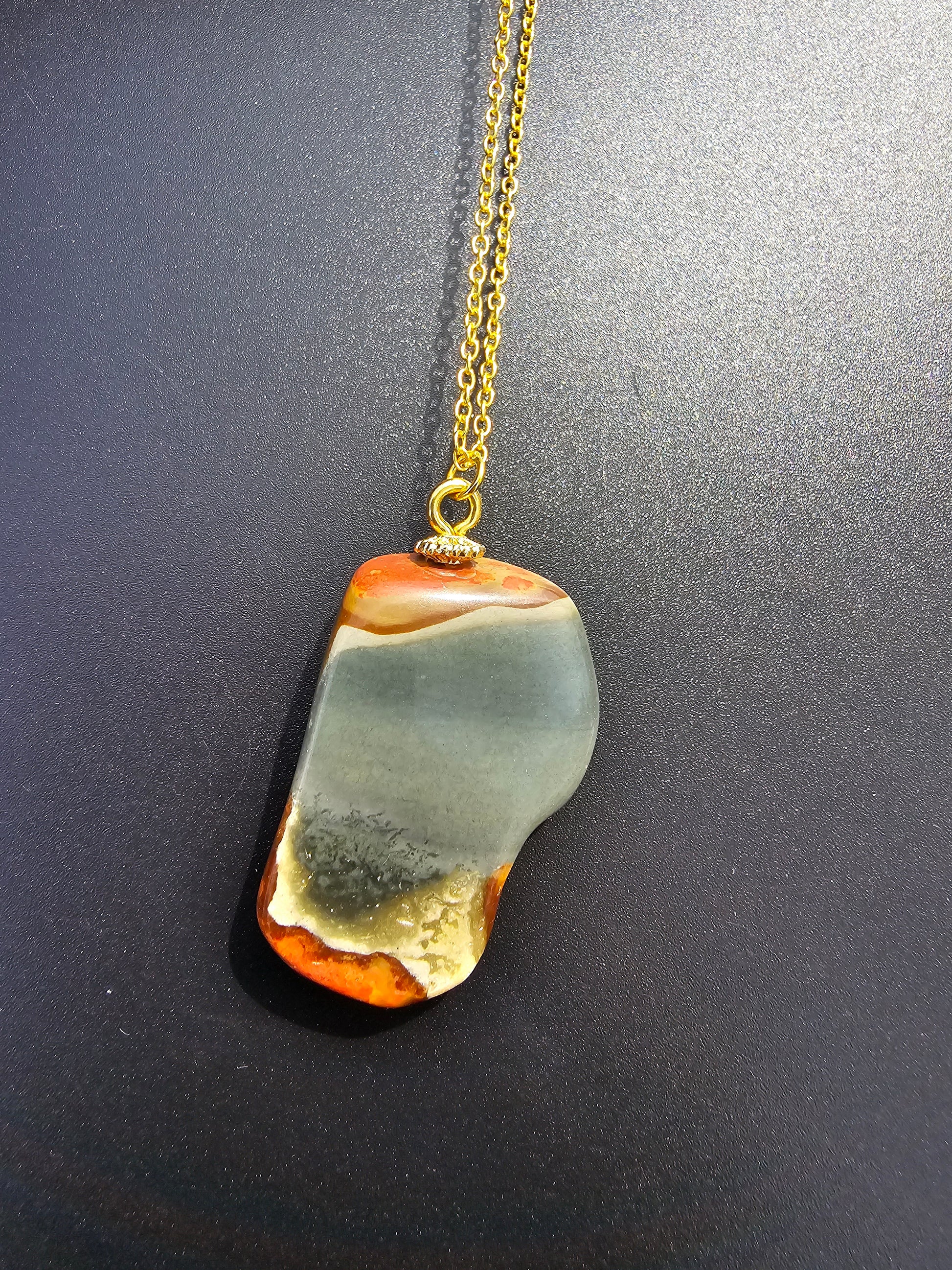 This stunning desert jasper pendant is a one-of-a-kind piece that will elevate any outfit. With intricate patterns and warm earth tones, it adds a touch of natural beauty to your look. Pictured here on a dark background that accentuates its greys, and browns and other earth tones. Plated gold chain adorns this Pendant.