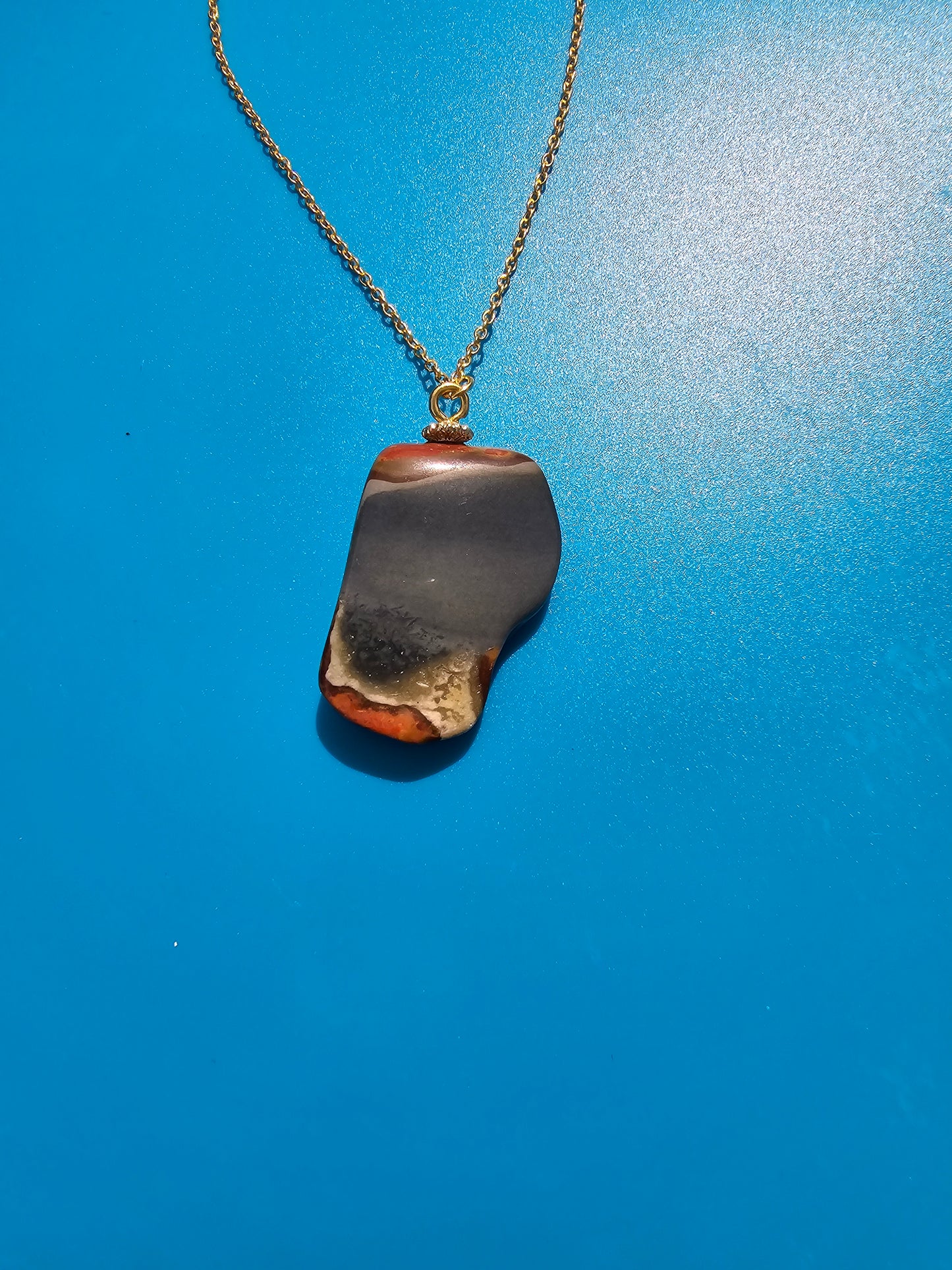 This stunning desert jasper pendant is a one-of-a-kind piece that will elevate any outfit. With intricate patterns and warm earth tones, it adds a touch of natural beauty to your look. Pictured here on a blue background that accentuates its greys, and browns and other earth tones. Plated gold chain adorns this Pendant.