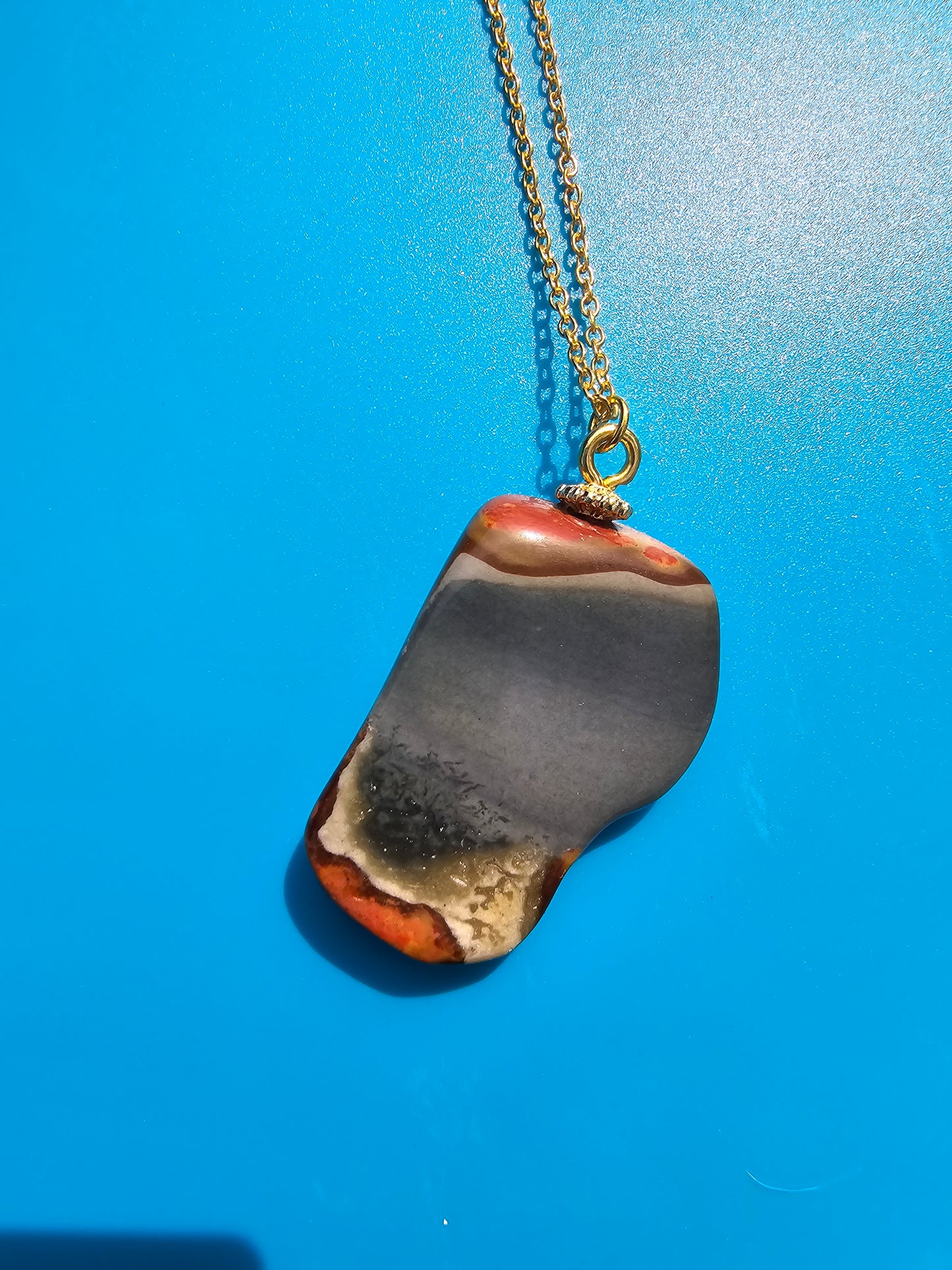 This stunning desert jasper pendant is a one-of-a-kind piece that will elevate any outfit. With intricate patterns and warm earth tones, it adds a touch of natural beauty to your look. Pictured here on a blue background that accentuates its greys, and browns and other earth tones. Plated gold chain adorns this Pendant.