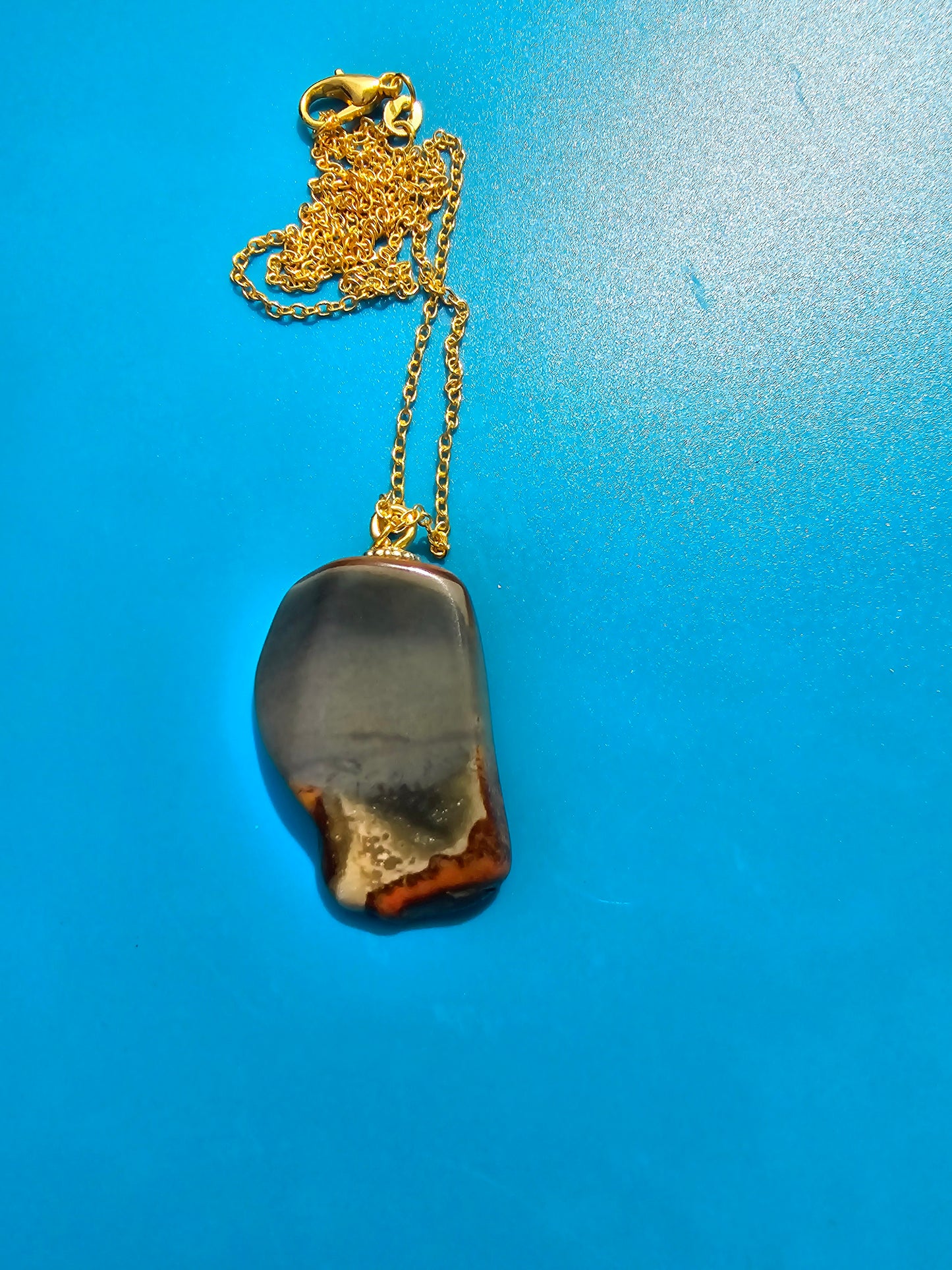 This stunning desert jasper pendant is a one-of-a-kind piece that will elevate any outfit. With intricate patterns and warm earth tones, it adds a touch of natural beauty to your look. Pictured here on a blue background that accentuates its greys, and browns and other earth tones. Plated gold chain adorns this Pendant.