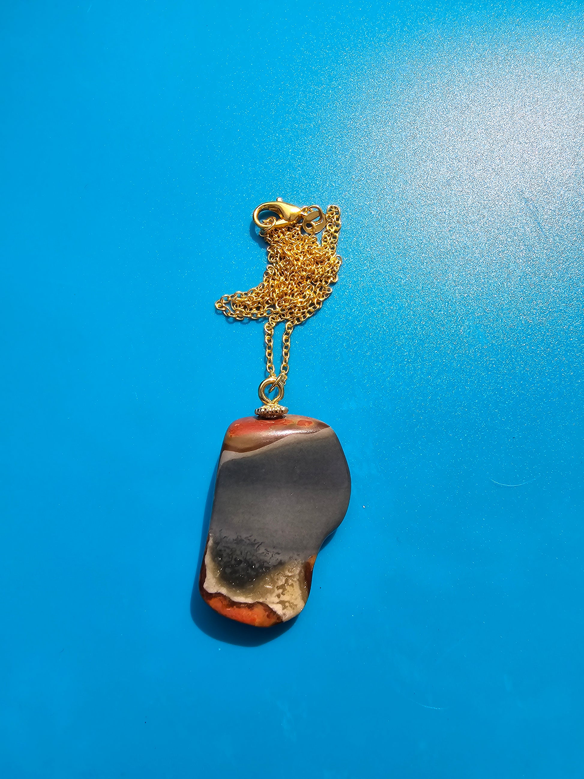 This stunning desert jasper pendant is a one-of-a-kind piece that will elevate any outfit. With intricate patterns and warm earth tones, it adds a touch of natural beauty to your look. Pictured here on a blue background that accentuates its greys, and browns and other earth tones. Plated gold chain adorns this Pendant.