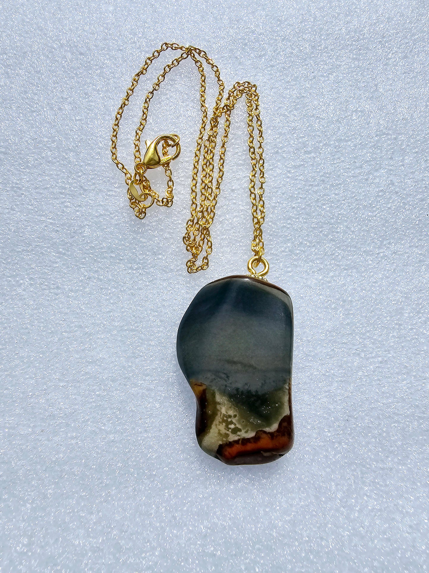 This stunning desert jasper pendant is a one-of-a-kind piece that will elevate any outfit. With intricate patterns and warm earth tones, it adds a touch of natural beauty to your look. Pictured here on a white background that accentuates its greys, and browns and other earth tones. Plated gold chain adorns this Pendant.