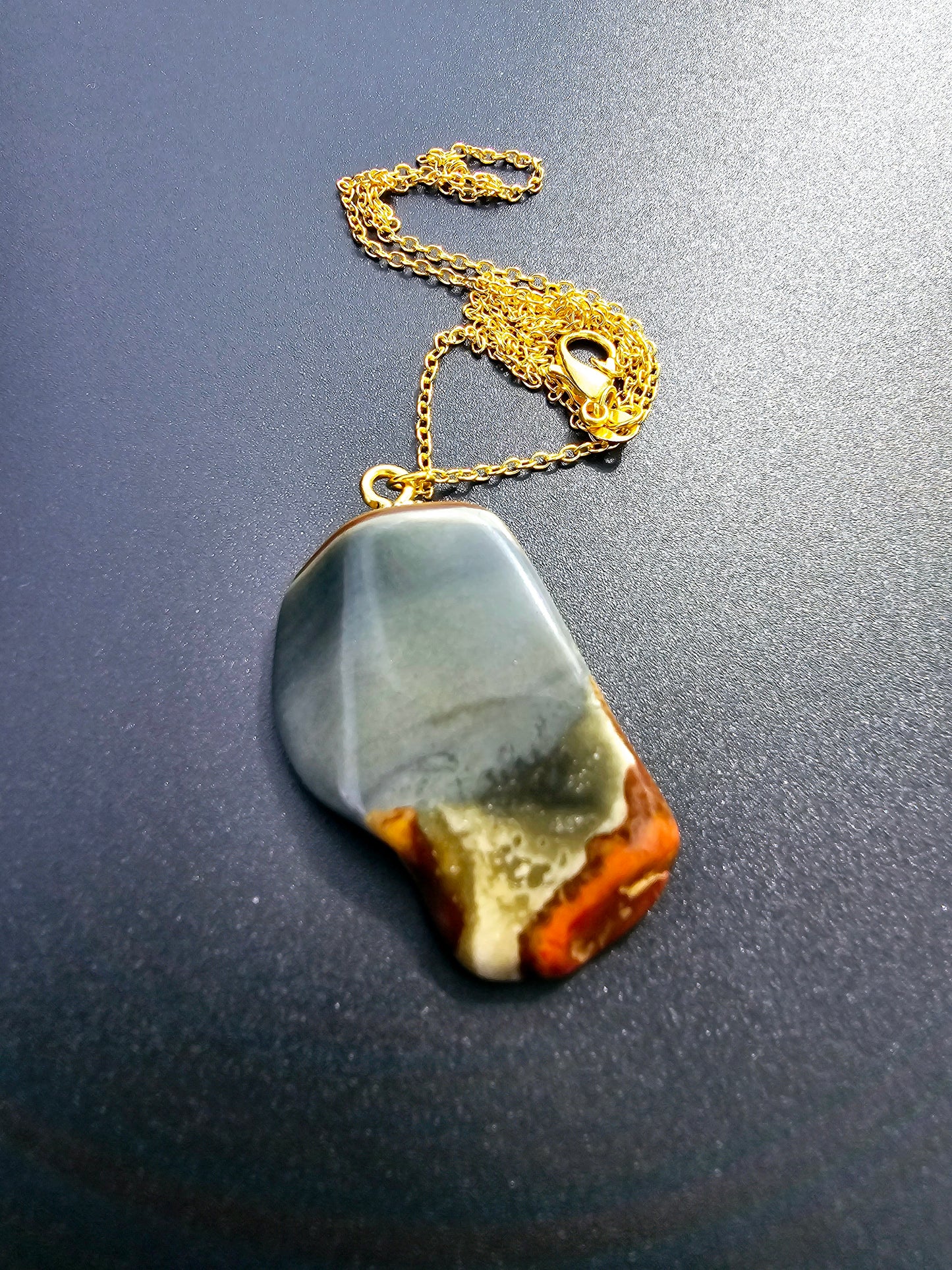 This stunning desert jasper pendant is a one-of-a-kind piece that will elevate any outfit. With intricate patterns and warm earth tones, it adds a touch of natural beauty to your look. Pictured here on a dark background that accentuates its greys, and browns and other earth tones. Plated gold chain adorns this Pendant.