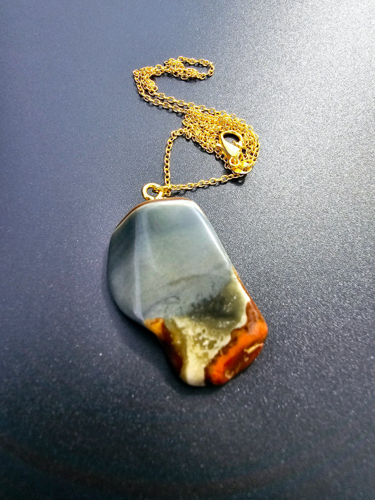 This stunning desert jasper pendant is a one-of-a-kind piece that will elevate any outfit. With intricate patterns and warm earth tones, it adds a touch of natural beauty to your look. Pictured here on a dark background that accentuates its greys, and browns and other earth tones. Plated gold chain adorns this Pendant.