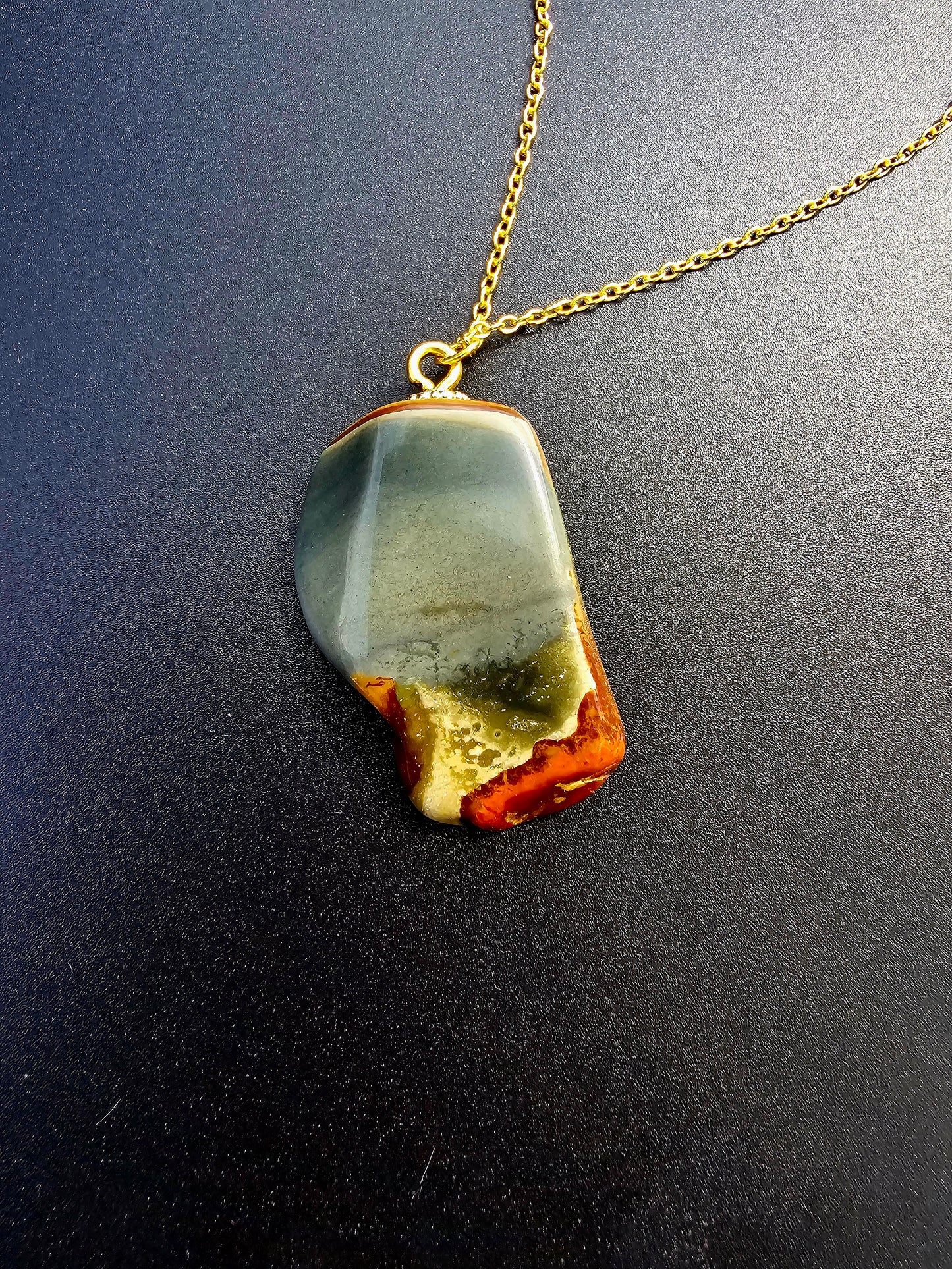 This stunning desert jasper pendant is a one-of-a-kind piece that will elevate any outfit. With intricate patterns and warm earth tones, it adds a touch of natural beauty to your look. Pictured here on a dark background that accentuates its greys, and browns and other earth tones. Plated gold chain adorns this Pendant.