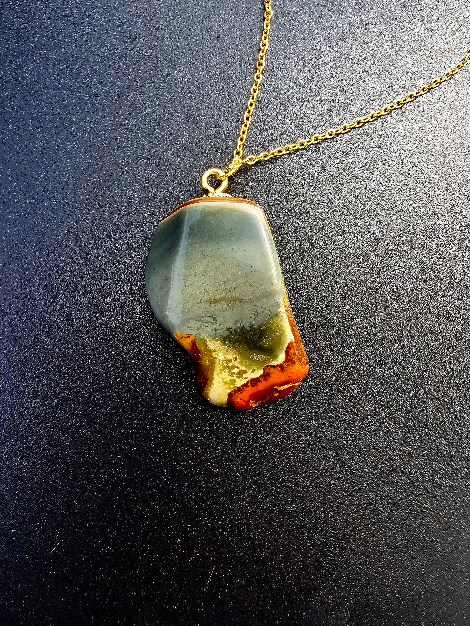 This stunning desert jasper pendant is a one-of-a-kind piece that will elevate any outfit. With intricate patterns and warm earth tones, it adds a touch of natural beauty to your look. Pictured here on a dark background that accentuates its greys, and browns and other earth tones. Plated gold chain adorns this Pendant.