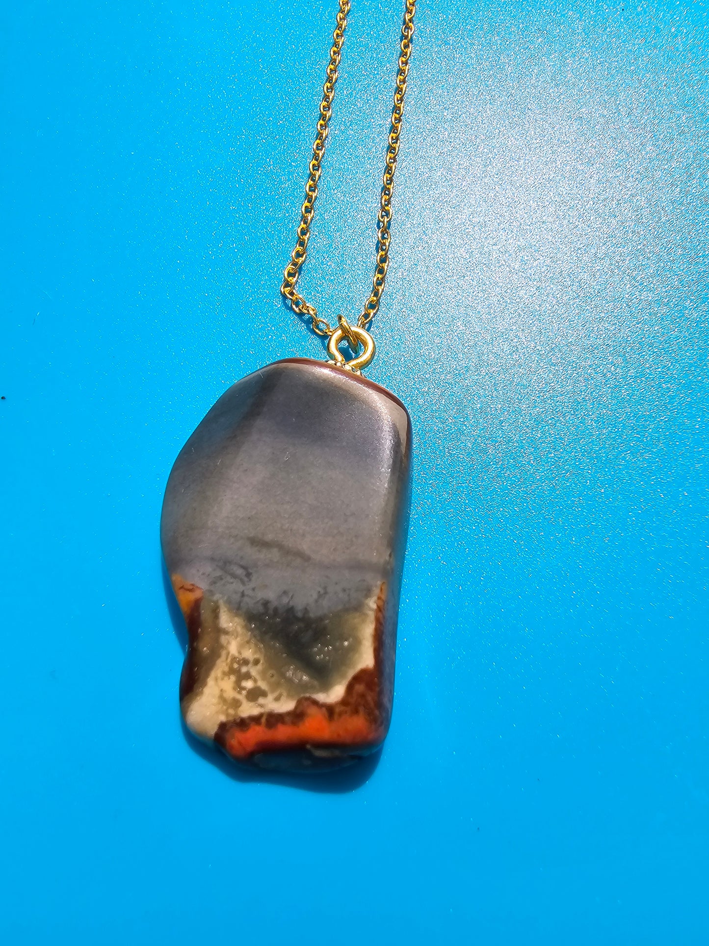 This stunning desert jasper pendant is a one-of-a-kind piece that will elevate any outfit. With intricate patterns and warm earth tones, it adds a touch of natural beauty to your look. Pictured here on a blue background that accentuates its greys, and browns and other earth tones. Plated gold chain adorns this Pendant.