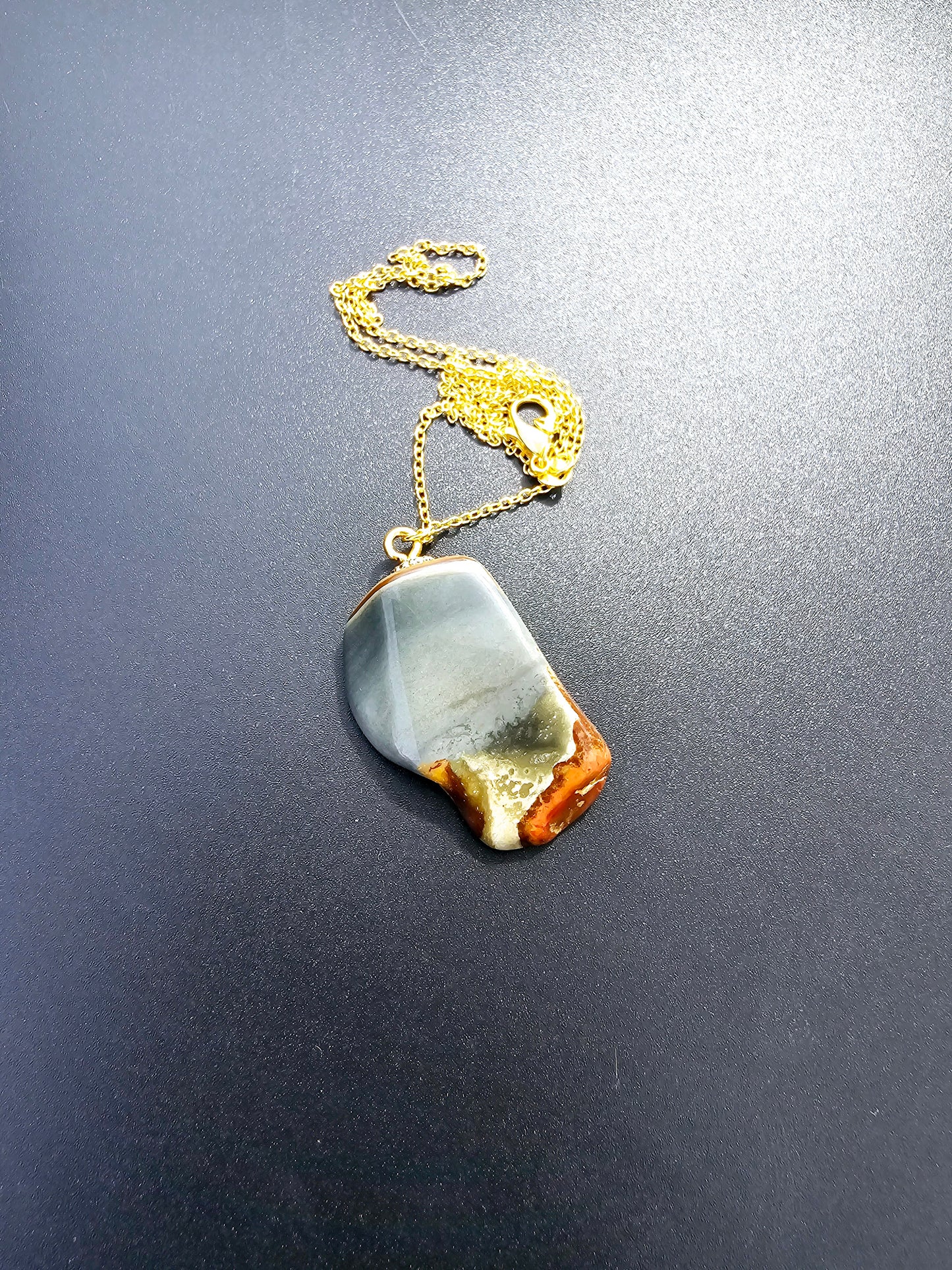 This stunning desert jasper pendant is a one-of-a-kind piece that will elevate any outfit. With intricate patterns and warm earth tones, it adds a touch of natural beauty to your look. Pictured here on a dark background that accentuates its greys, and browns and other earth tones. Plated gold chain adorns this Pendant.