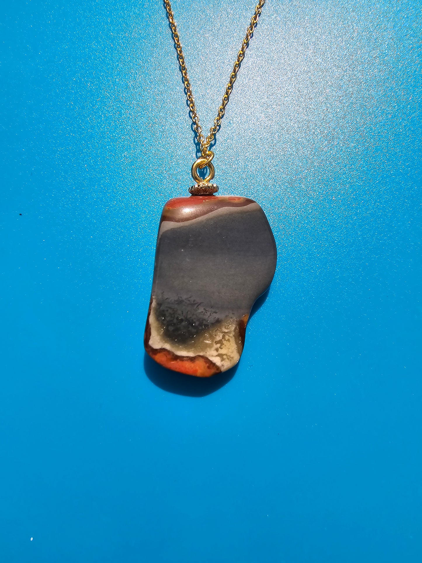 This stunning desert jasper pendant is a one-of-a-kind piece that will elevate any outfit. With intricate patterns and warm earth tones, it adds a touch of natural beauty to your look. Pictured here on a blue background that accentuates its greys, and browns and other earth tones. Plated gold chain adorns this Pendant.