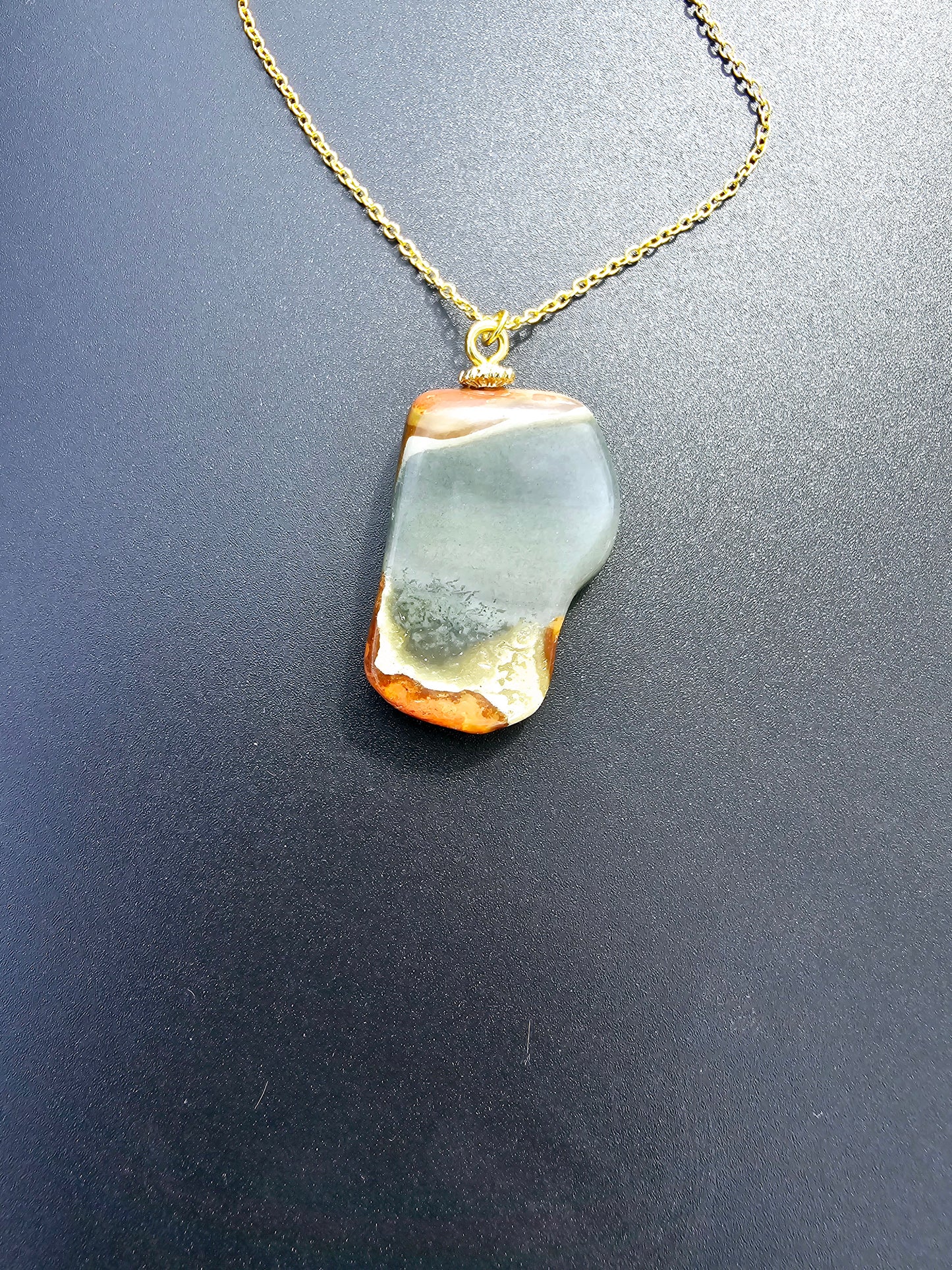 This stunning desert jasper pendant is a one-of-a-kind piece that will elevate any outfit. With intricate patterns and warm earth tones, it adds a touch of natural beauty to your look. Pictured here on a dark background that accentuates its greys, and browns and other earth tones. Plated gold chain adorns this Pendant.