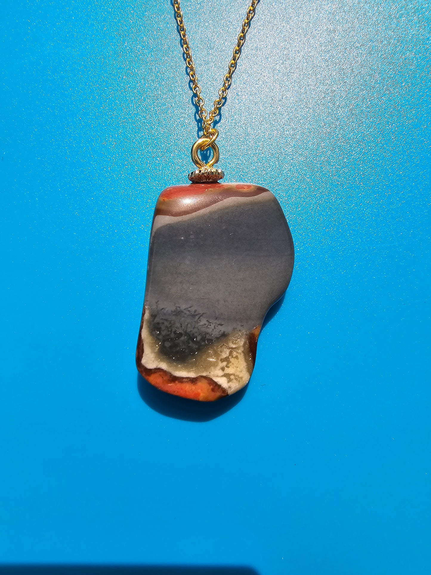 This stunning desert jasper pendant is a one-of-a-kind piece that will elevate any outfit. With intricate patterns and warm earth tones, it adds a touch of natural beauty to your look. Pictured here on a blue background that accentuates its greys, and browns and other earth tones. Plated gold chain adorns this Pendant.