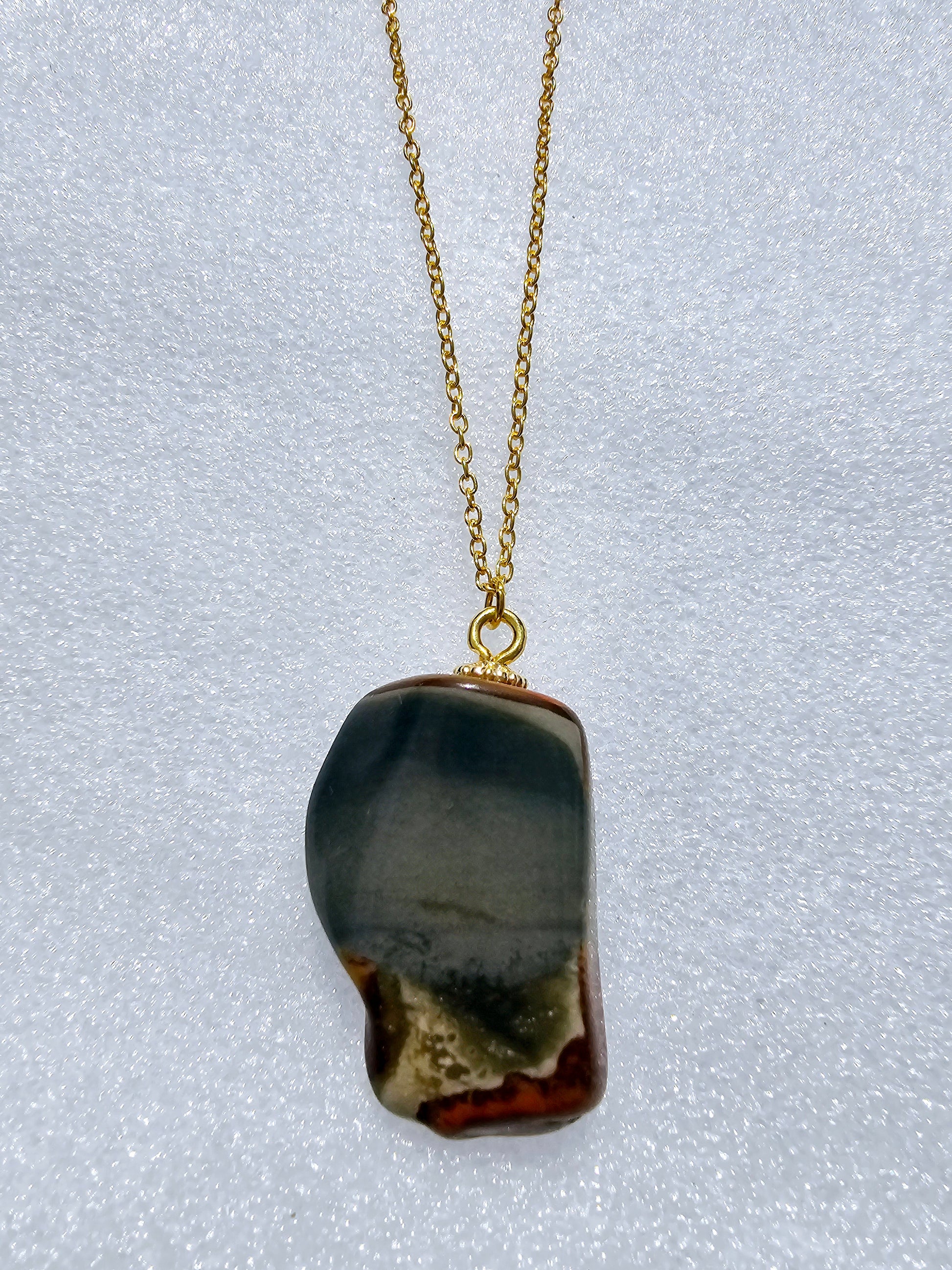 This stunning desert jasper pendant is a one-of-a-kind piece that will elevate any outfit. With intricate patterns and warm earth tones, it adds a touch of natural beauty to your look. Pictured here on a white background that accentuates its greys, and browns and other earth tones. Plated gold chain adorns this Pendant.