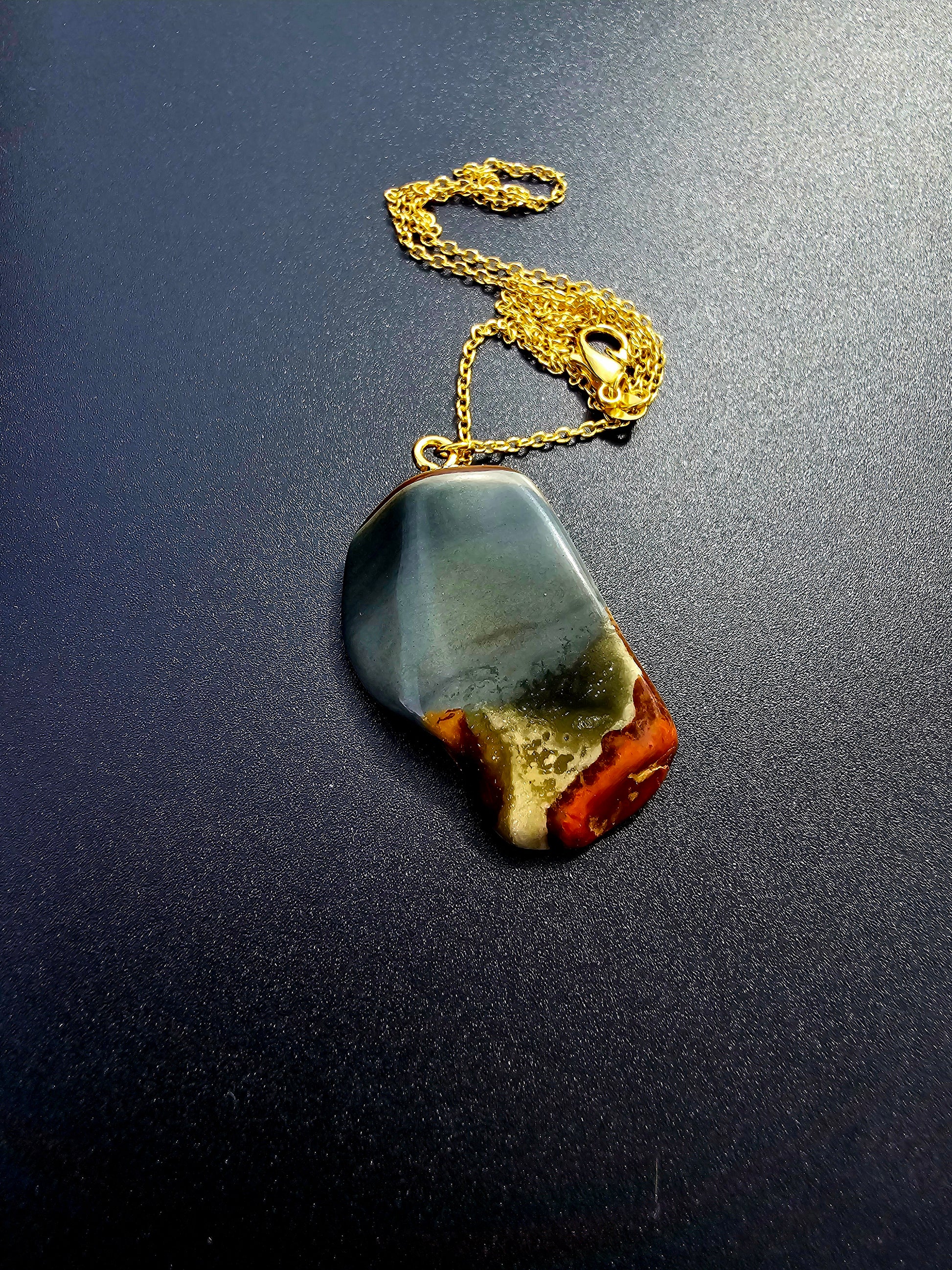 This stunning desert jasper pendant is a one-of-a-kind piece that will elevate any outfit. With intricate patterns and warm earth tones, it adds a touch of natural beauty to your look. Pictured here on a dark background that accentuates its greys, and browns and other earth tones. Plated gold chain adorns this Pendant.