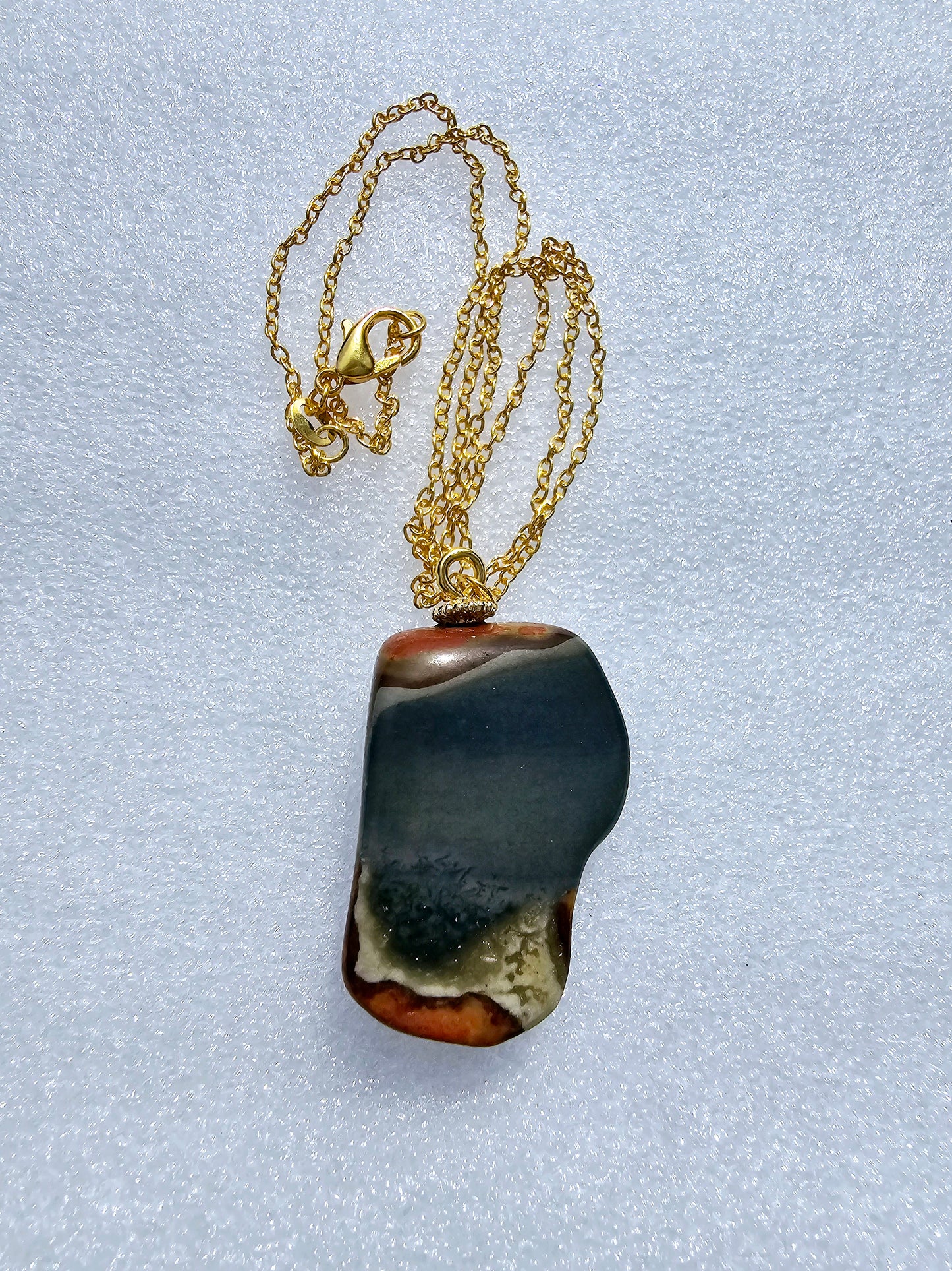 This stunning desert jasper pendant is a one-of-a-kind piece that will elevate any outfit. With intricate patterns and warm earth tones, it adds a touch of natural beauty to your look. Pictured here on a white background that accentuates its greys, and browns and other earth tones. Plated gold chain adorns this Pendant.